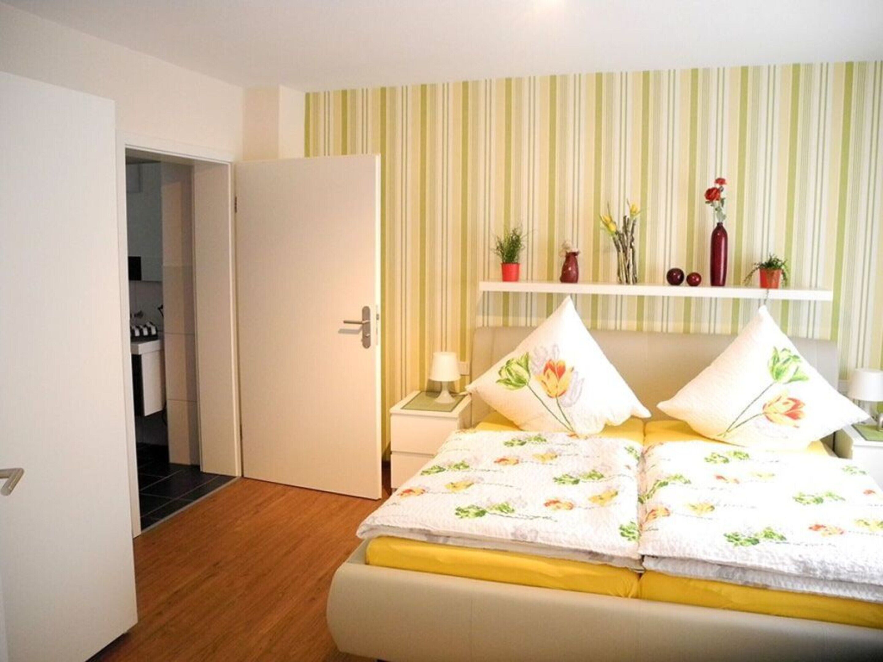 Ein Schlafzimmer mit Bett, Nachttisch, Ablagefläche und einem Regal mit Dekoration. Eine Tür führt in einen anderen Raum.