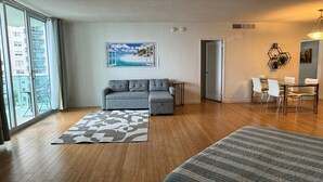 A spacious living area with a grey sofa, a patterned rug, a dining table with chairs, and a large window offering a view of buildings. - 7th FL spacious 1 bedroom  (Hollywood)