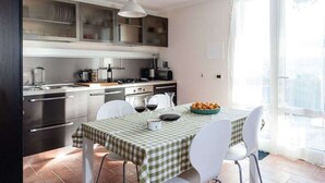 A modern kitchen with stainless steel appliances, a dining table with a checkered tablecloth, white chairs, and a large window providing natural light. - Casina di Savolano Comfortable Holiday Residence (Livorno)