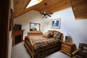 A bedroom with a sloped ceiling, a bed with plaid bedding, a nightstand with a lamp, a dresser, a mirror, and a fireplace.
