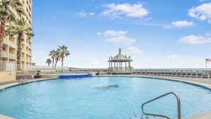 Une piscine avec un kiosque au centre, entourée de palmiers et d’un bâtiment d’hôtel.