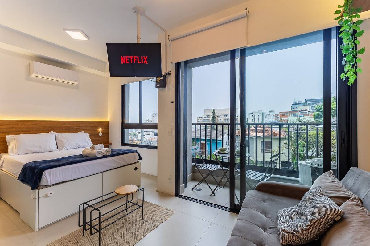 A modern bedroom with a bed, a TV mounted on the wall, a balcony with a table and chairs, and a city view.