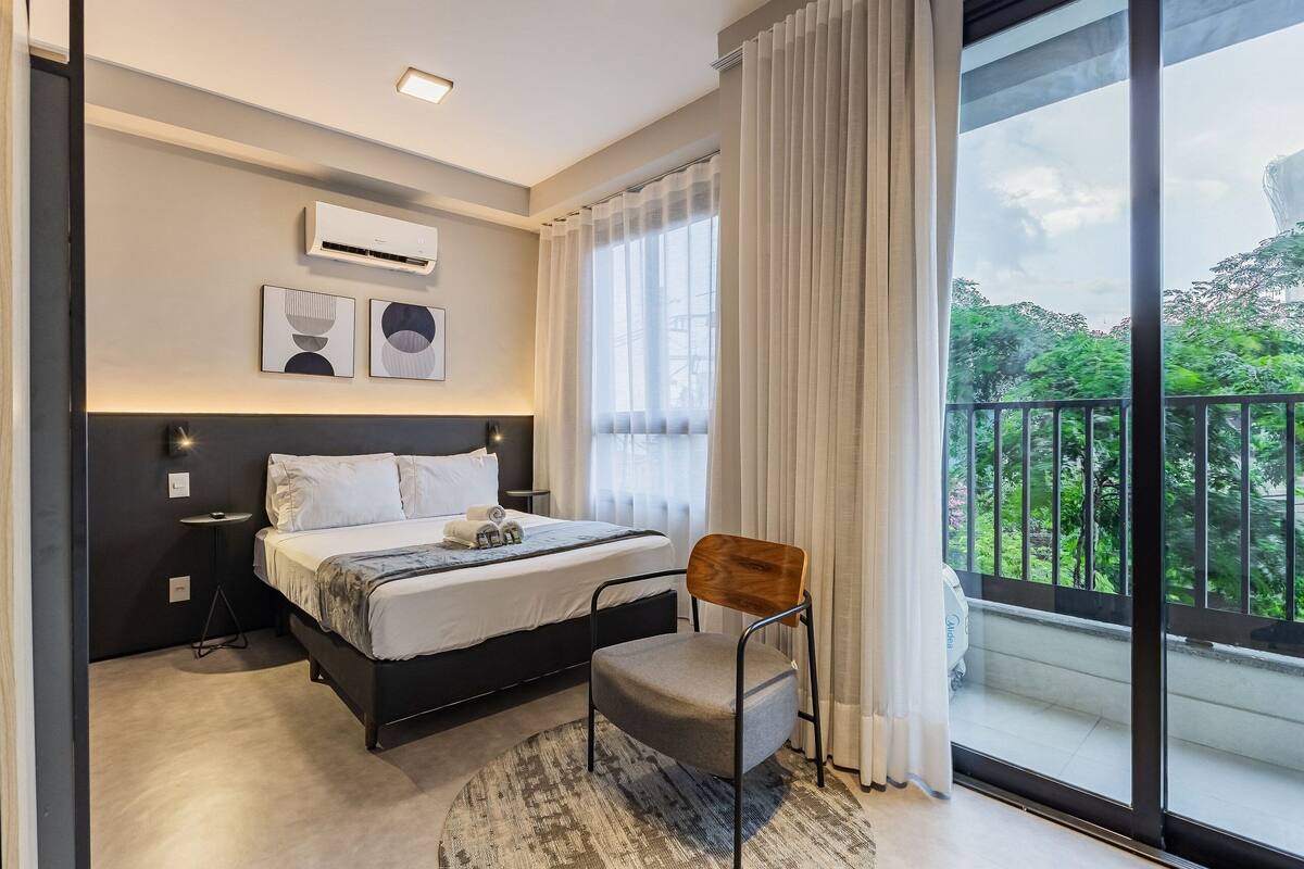 A modern hotel room with a bed, a chair, a small table, a window with curtains, and a balcony with a railing.