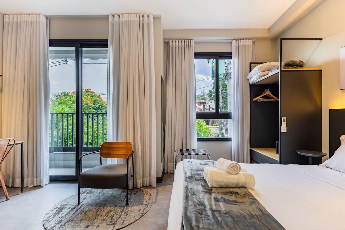 A hotel room with a bed, a chair, a small table, a wardrobe, and a view through a large window.
