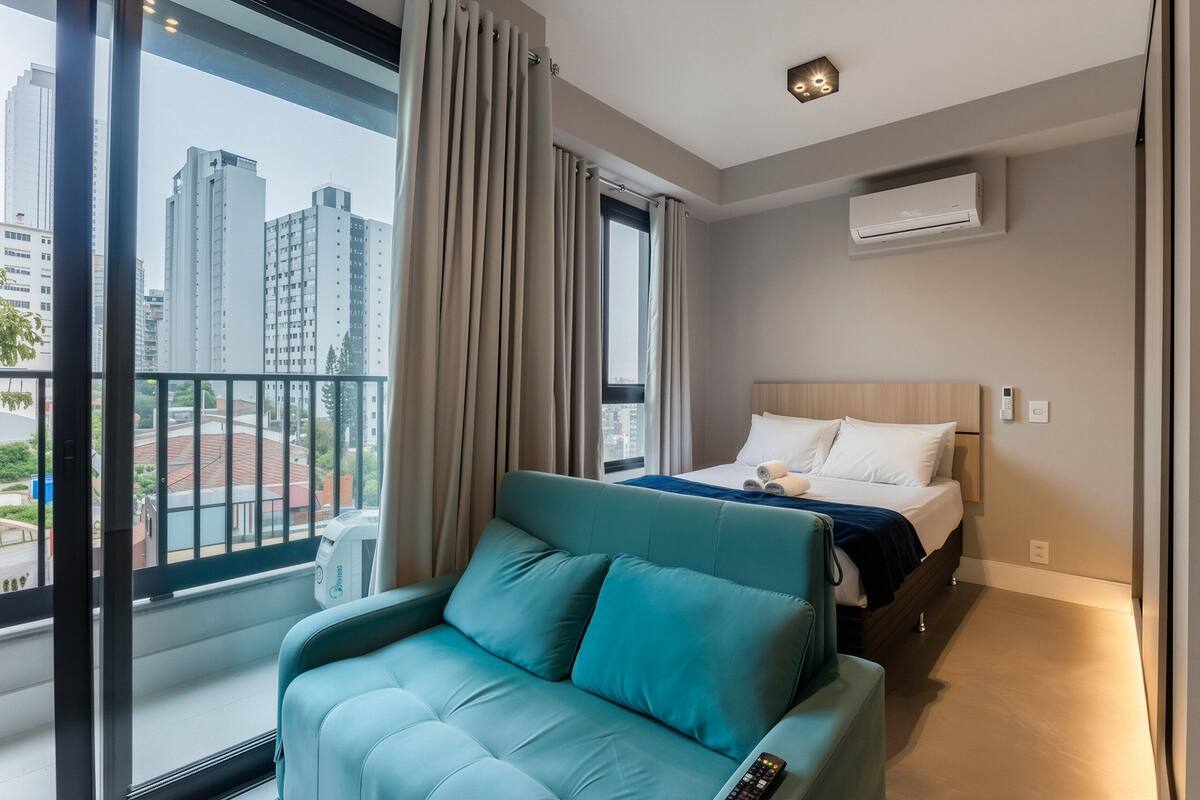 A hotel room with a balcony, a bed with white linens, a teal sofa, and an air conditioning unit on the wall.