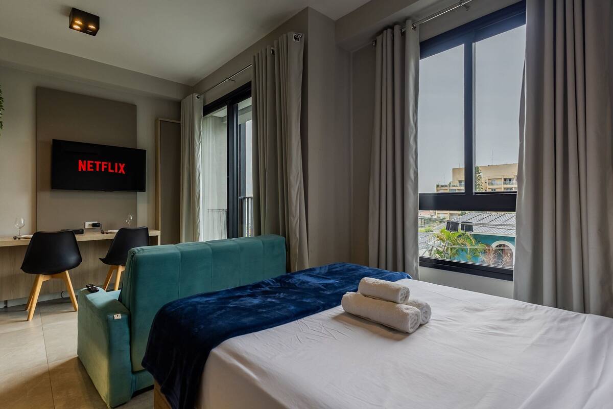 A hotel room with a bed, a desk, a chair, a TV displaying 'NETFLIX,' and a view of buildings through the window.