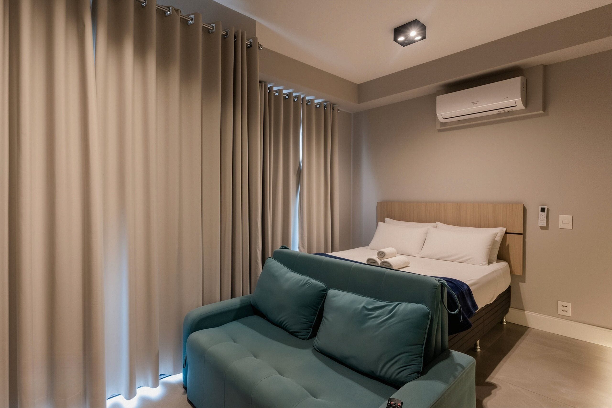 A modern hotel room with a bed, a sofa, curtains, an air conditioning unit, and a ceiling light.