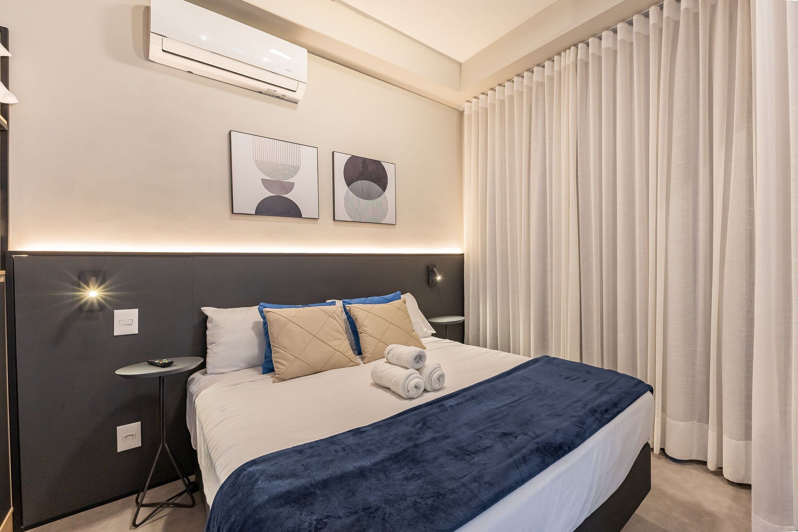 A modern hotel room with a large bed, a bedside table, a wall-mounted air conditioner, and a window with sheer curtains.