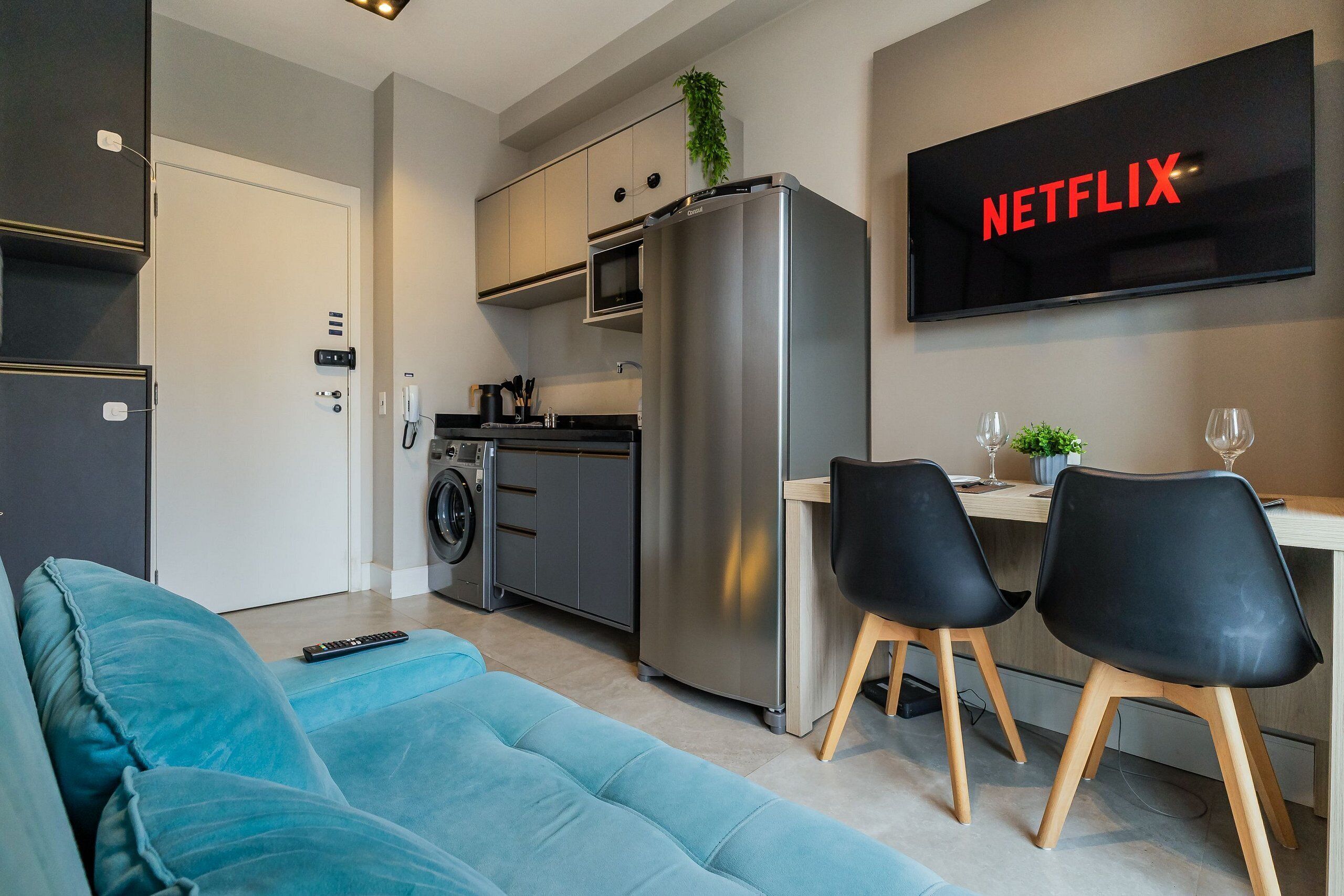 Eine moderne Küche mit einem Edelstahl-Kühlschrank, einer Mikrowelle und einem Backofen, eine Essecke mit zwei Stühlen, einem Sofa und einem Fernseher, der 'NETFLIX' anzeigt.