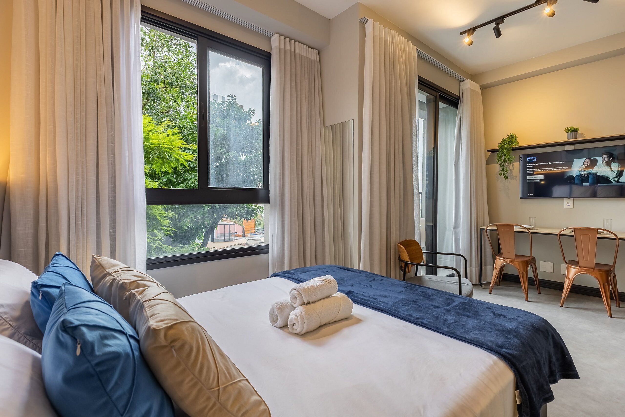 Une chambre d’hôtel avec un lit, un bureau, deux chaises, une télévision et une fenêtre avec des rideaux.