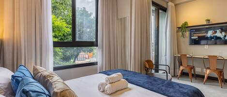 Une chambre d’hôtel avec un lit, un bureau, deux chaises, une télévision et une fenêtre avec des rideaux.