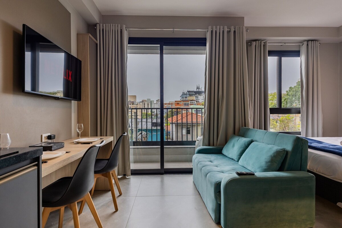 Habitación de hotel moderna con zona de comedor, sofá, televisor y balcón con vistas a la ciudad.