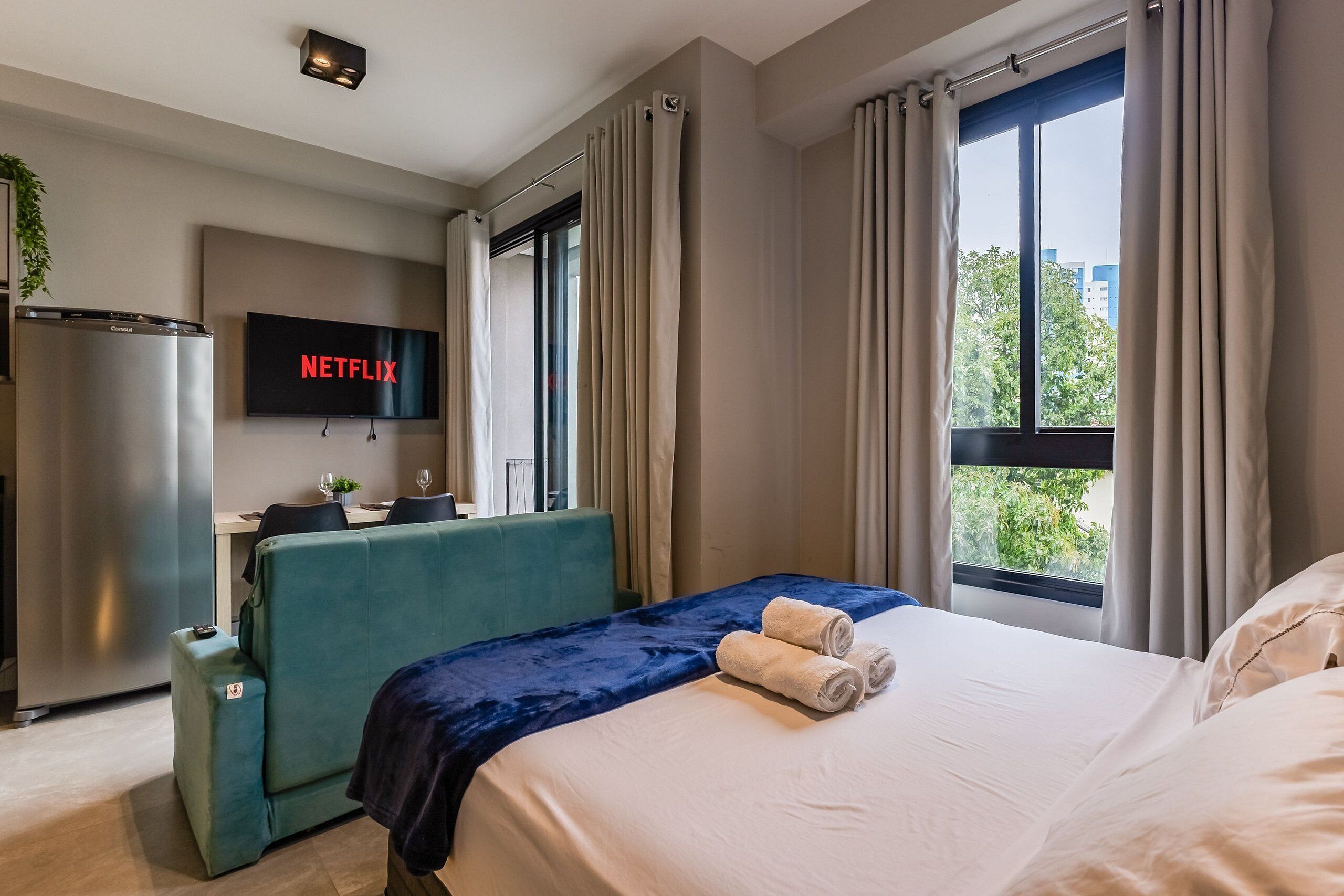 Una habitació d'hotel amb un llit, un televisor que mostra 'NETFLIX', un sofà, una taula petita amb una planta i una finestra amb cortines.