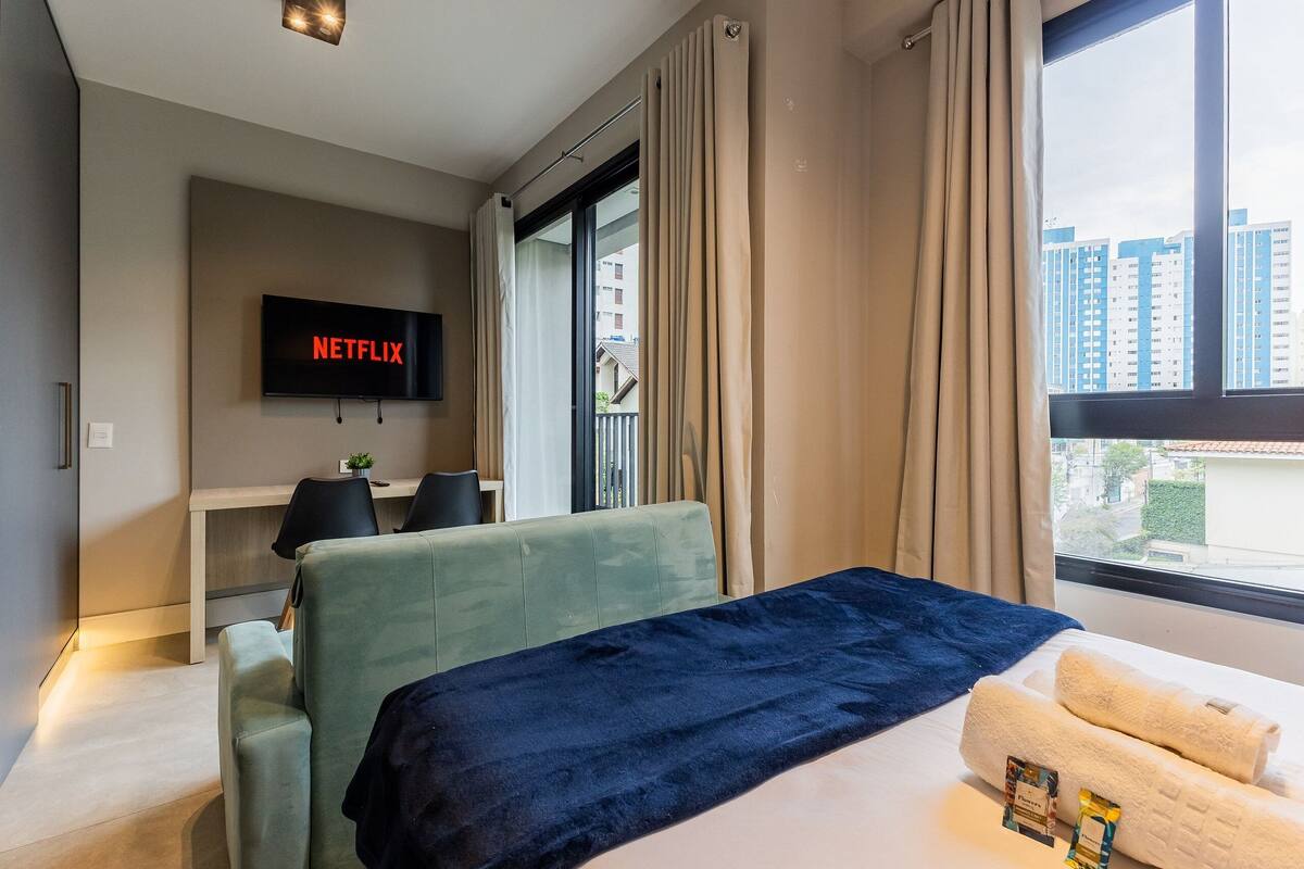 A hotel room with a bed, a desk, a chair, a TV displaying 'NETFLIX,' and a view of buildings through the window.