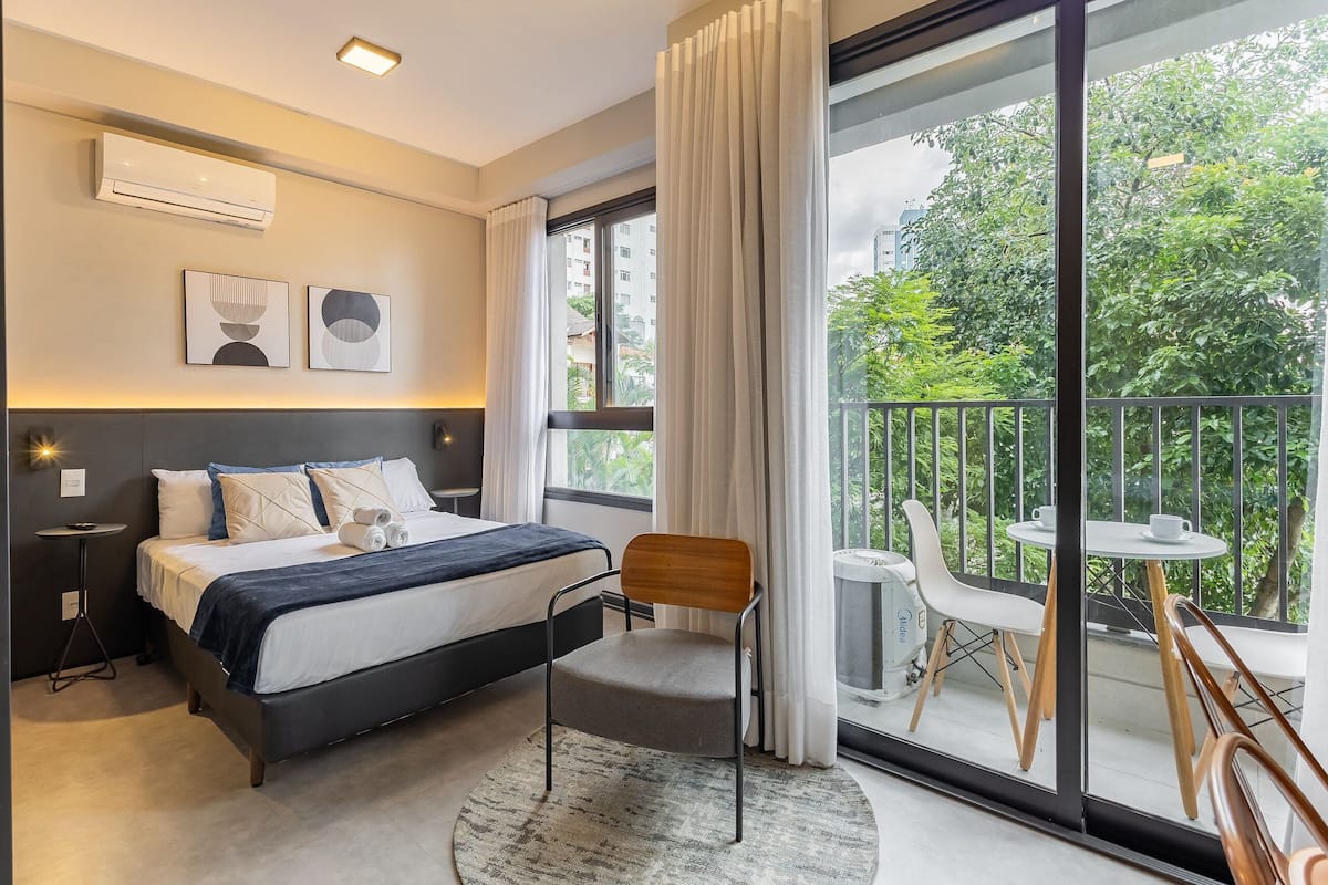 A modern hotel room with a bed, a chair, a small table, and a balcony with a view of greenery.
