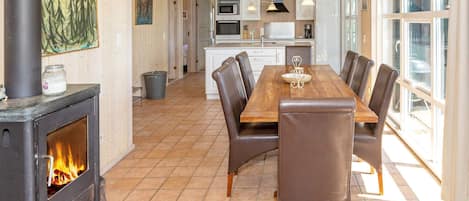 A spacious dining area with a wooden table and leather chairs, a kitchen in the background, and a wood-burning stove.