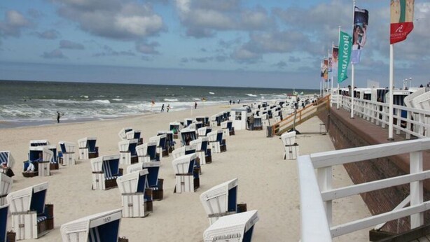 Ein Strand mit Reihen aus weißen und blauen Liegestühlen und Sonnenschirmen, einer weißen Brüstung und Blick auf das Meer.