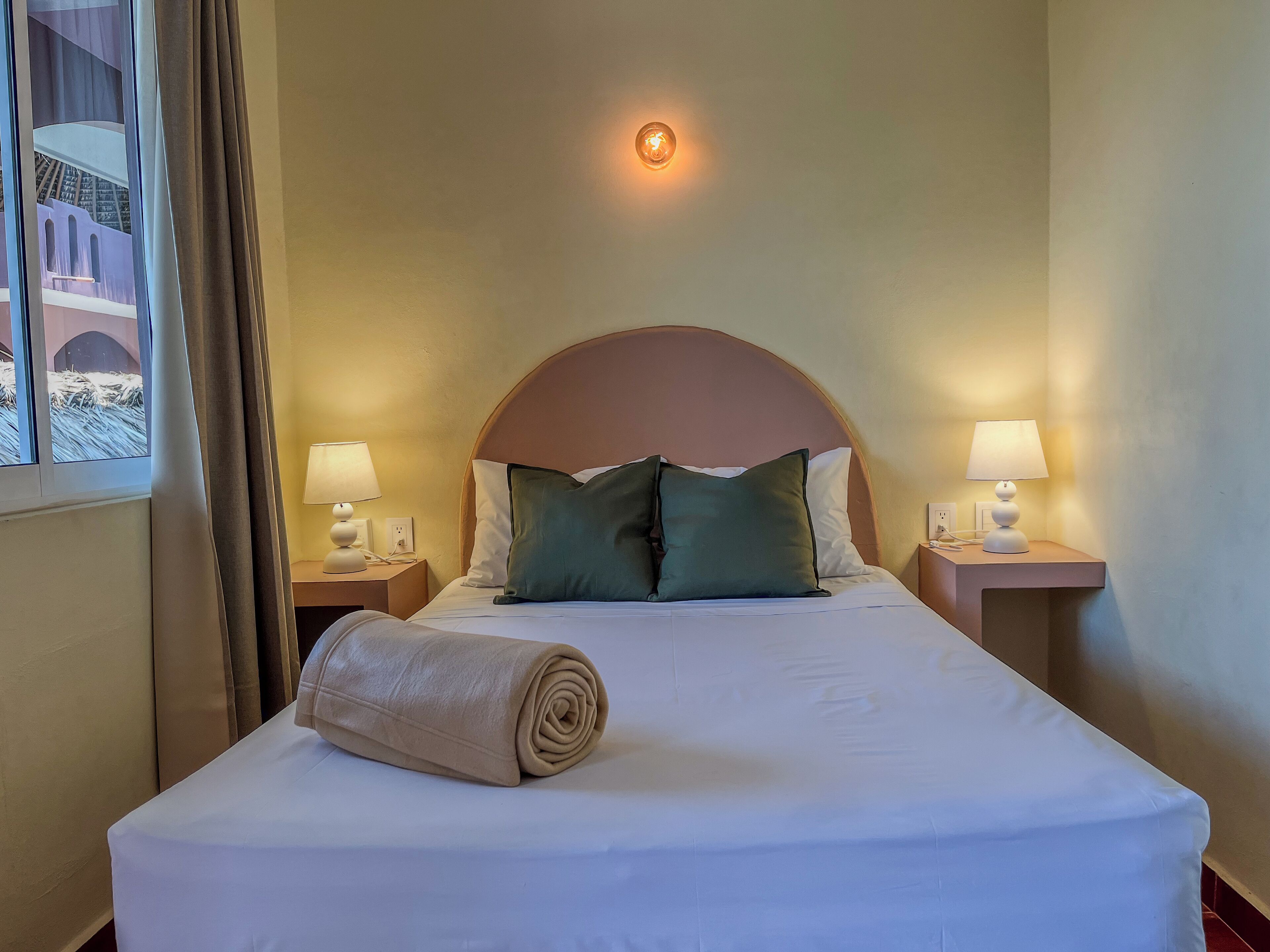 A hotel room with a bed, two bedside lamps, a window with curtains, and a view of a building outside.