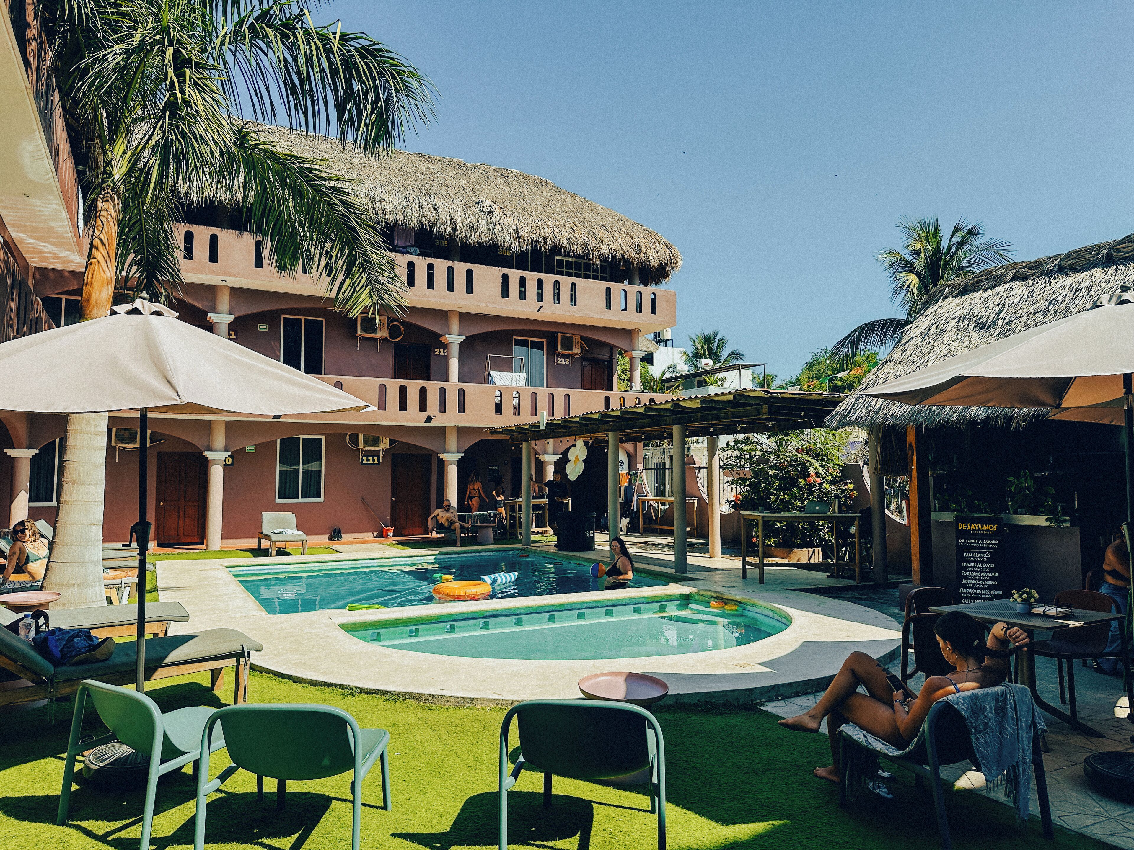 A pool area with lounge chairs, a thatched-roof building, and a restaurant.