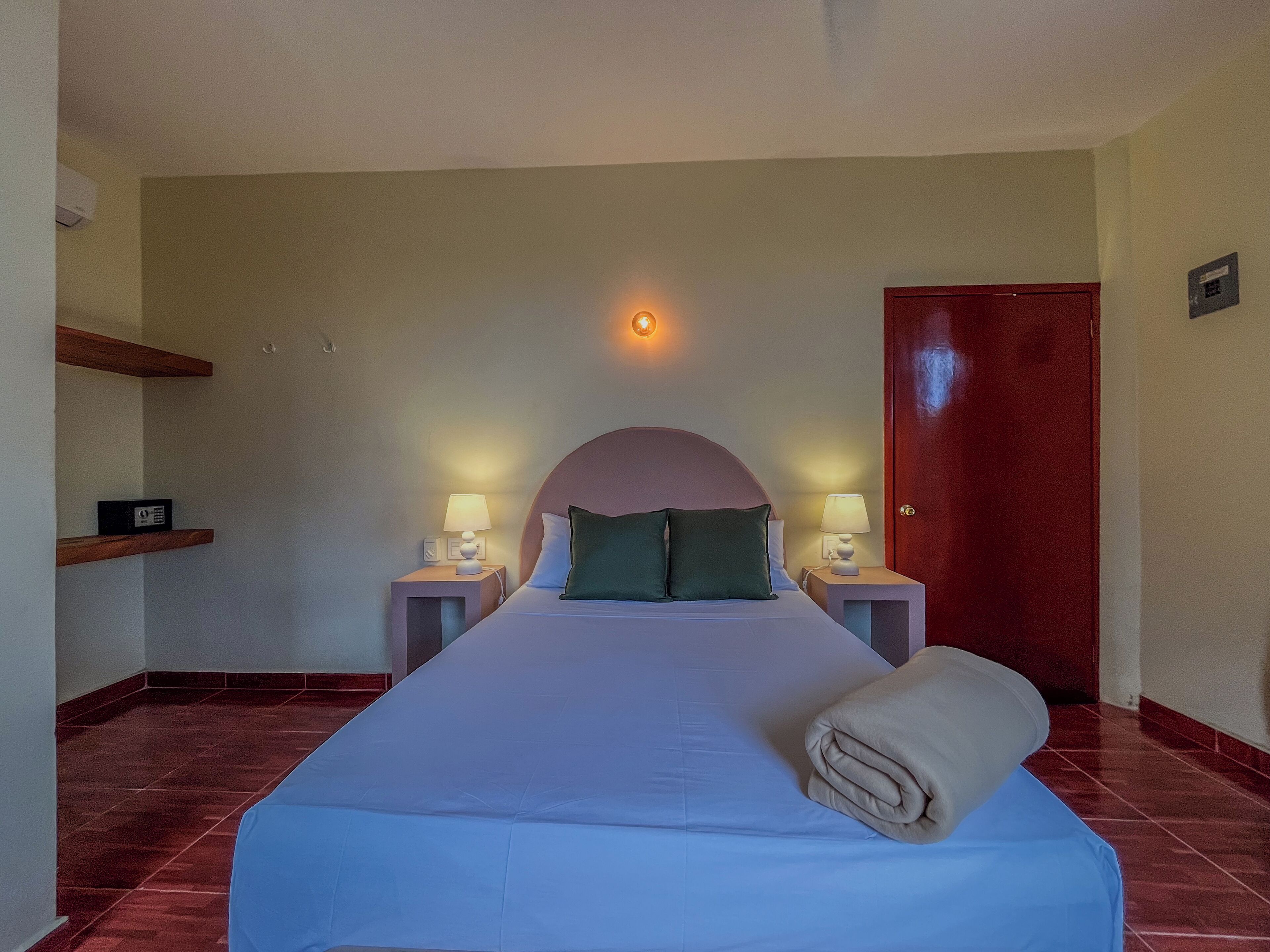 Une chambre d’hôtel avec un grand lit, deux tables de chevet avec des lampes, une porte rouge et une petite étagère avec une radio.