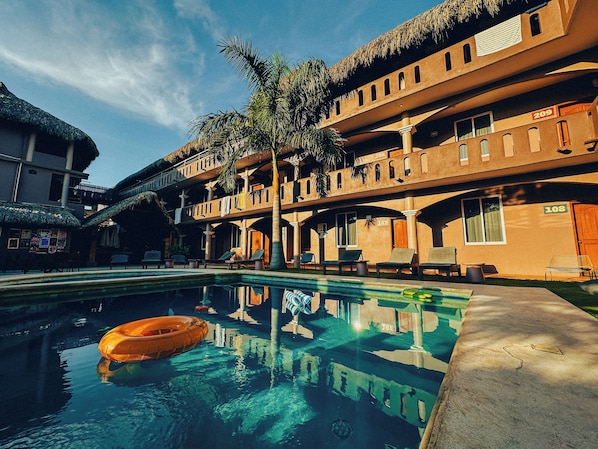 Ein Pool mit Liegestühlen und einem Schwimmreifen, umgeben von einem Gebäude mit mehreren Balkonen und Palmen.