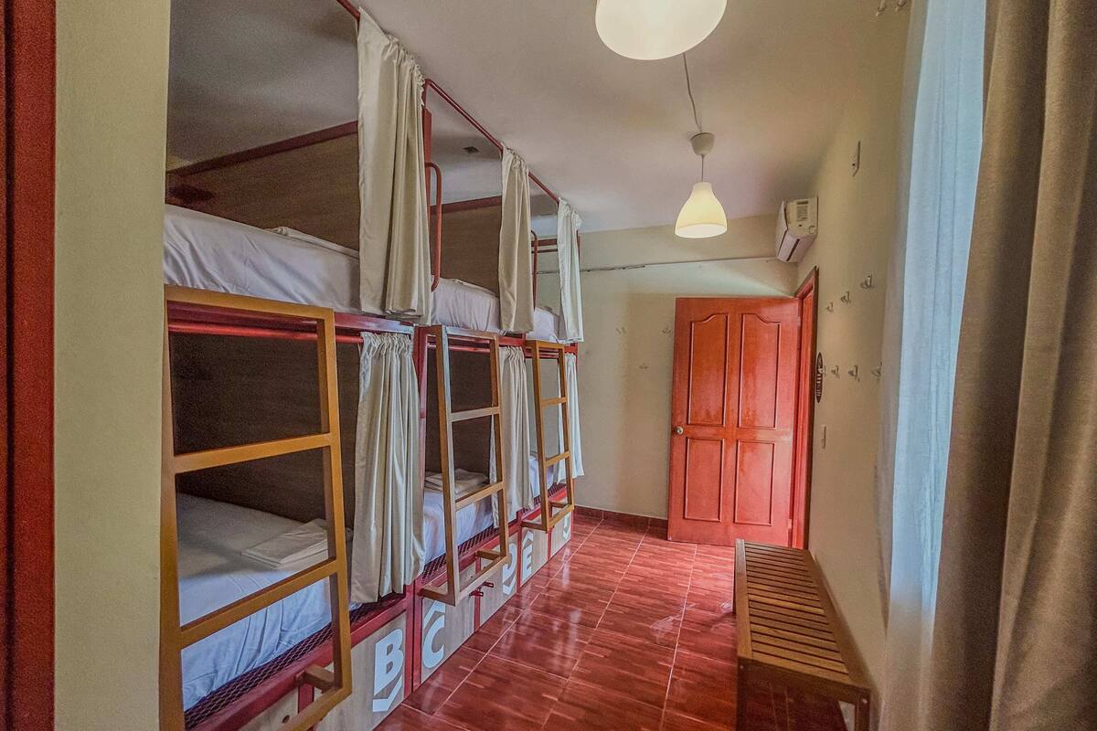 Asuntolan huone, jossa on kerrossängyt, puinen penkki, punainen ovi ja katosta roikkuva valaisin.
