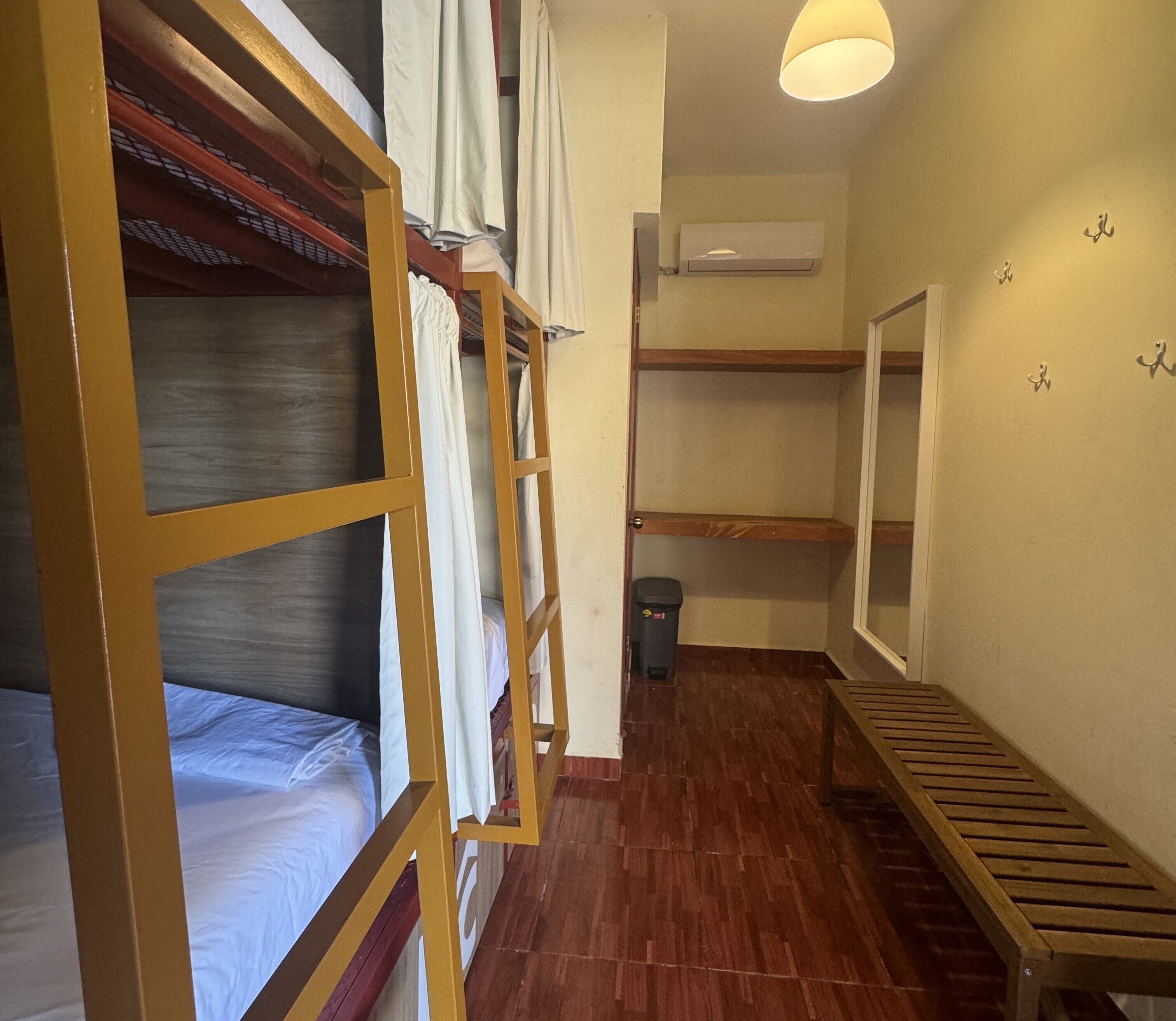 Ein Schlafsaalzimmer mit Etagenbetten, einer hölzernen Bank, einem Mülleimer und einer wandmontierten Klimaanlage.