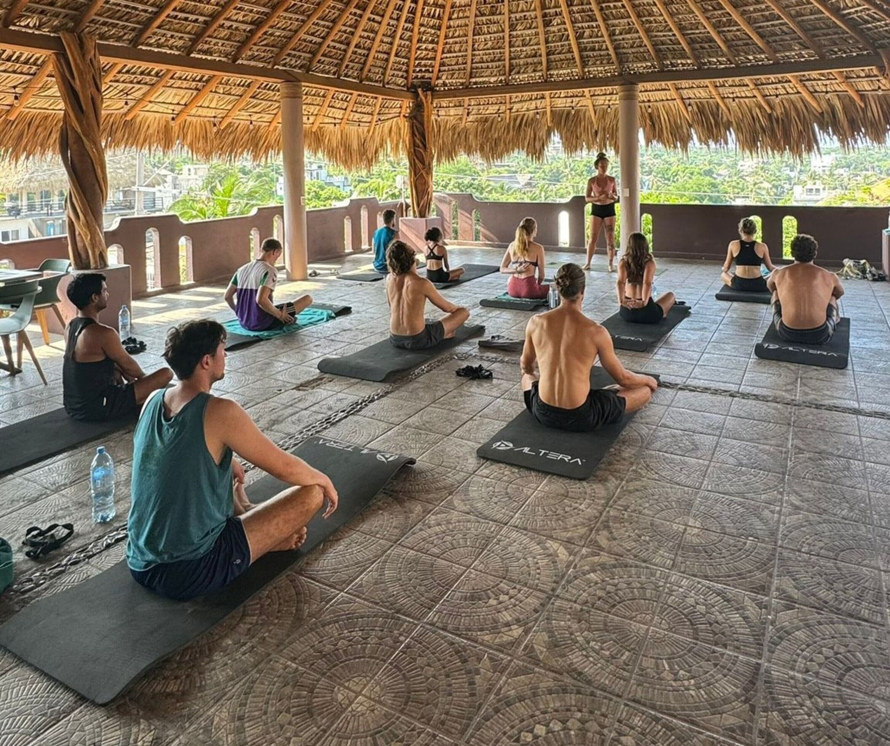 A group of people sitting on yoga mats in a covered area with a patterned floor.