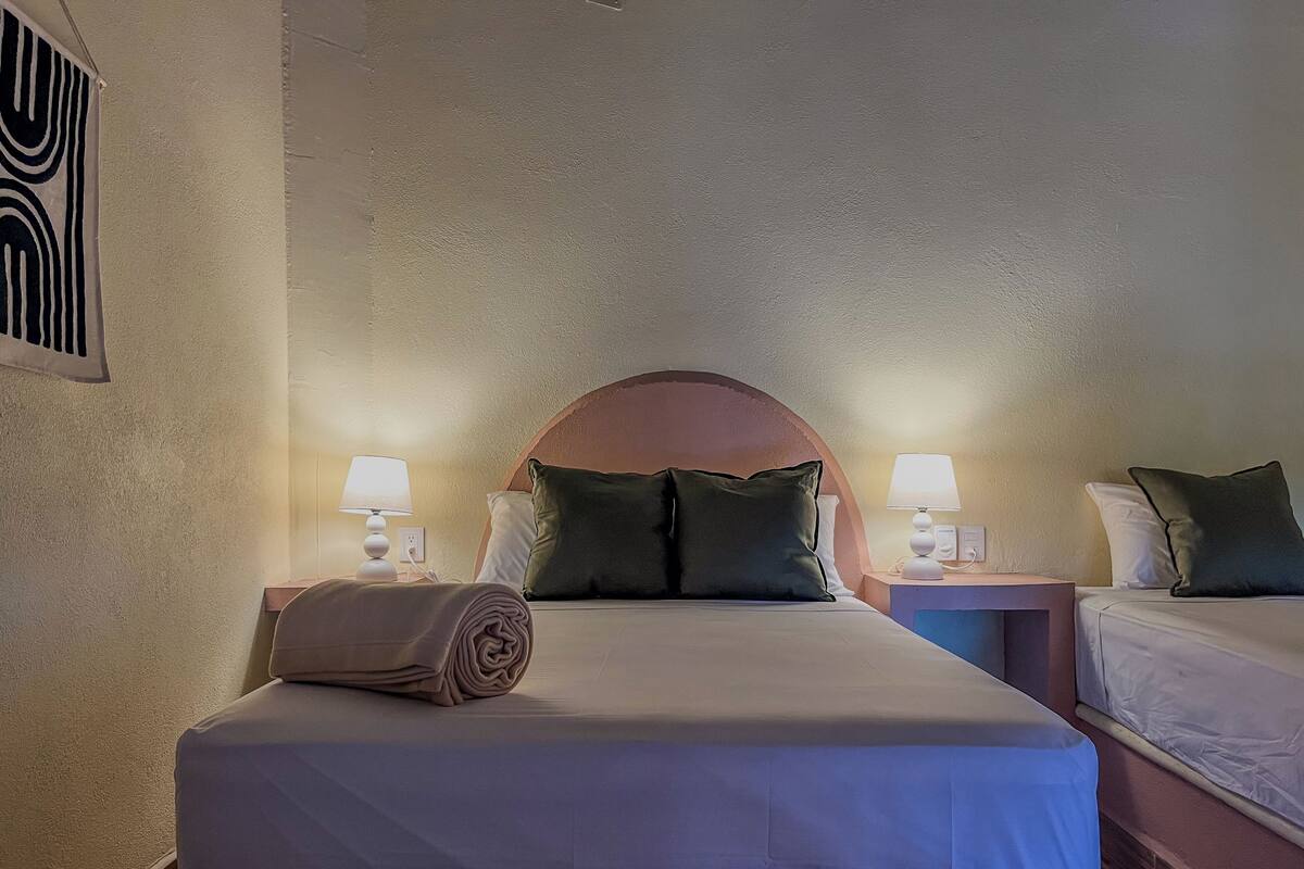 Hotellihuone, jossa on suuri sänky, kaksi yöpöytää valaisimineen, sängynpääty ja seinään asennettu ilmanvaihtoaukko.