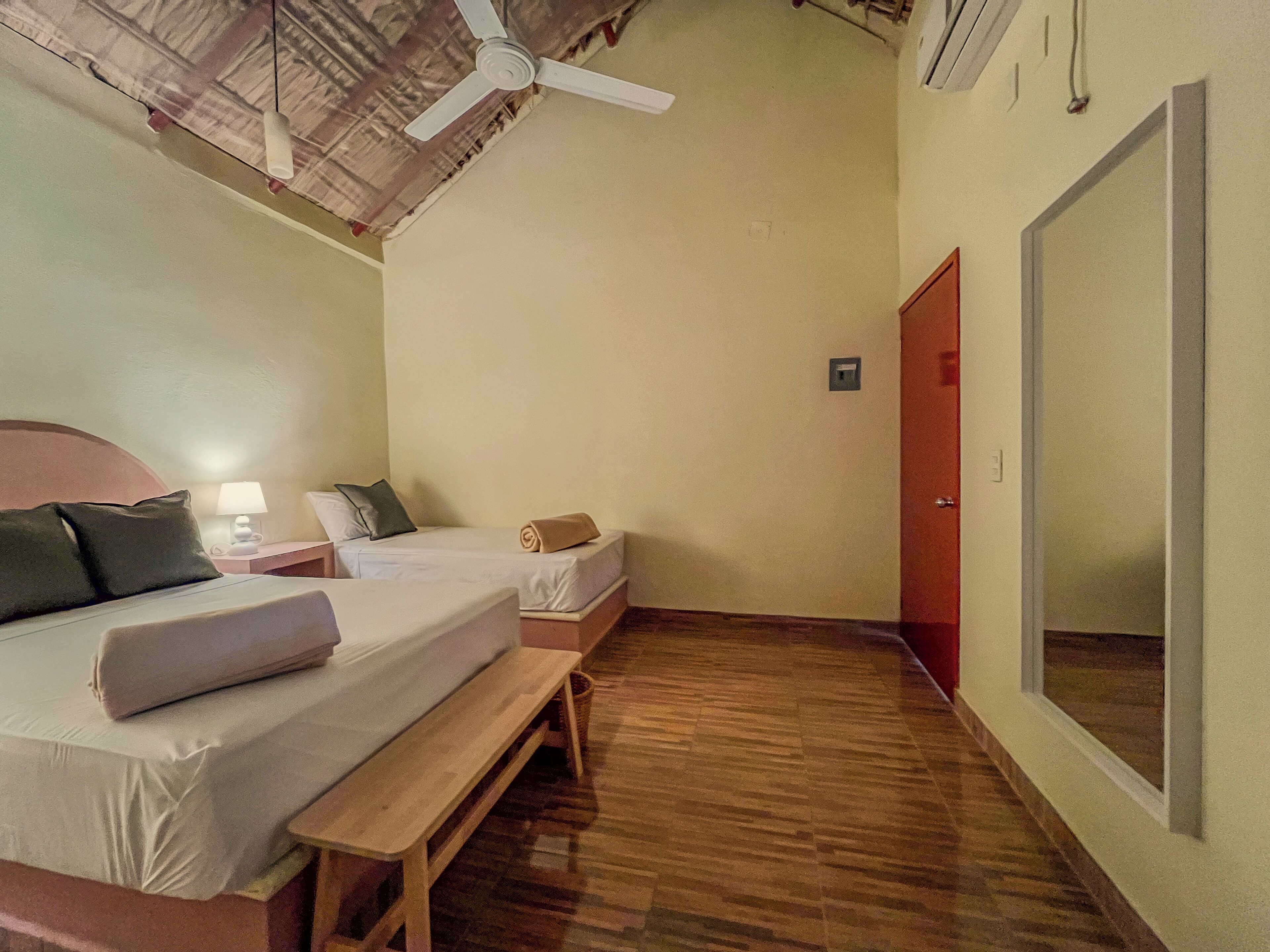 A room with two beds, a ceiling fan, a wooden bench, and a large mirror on the wall.