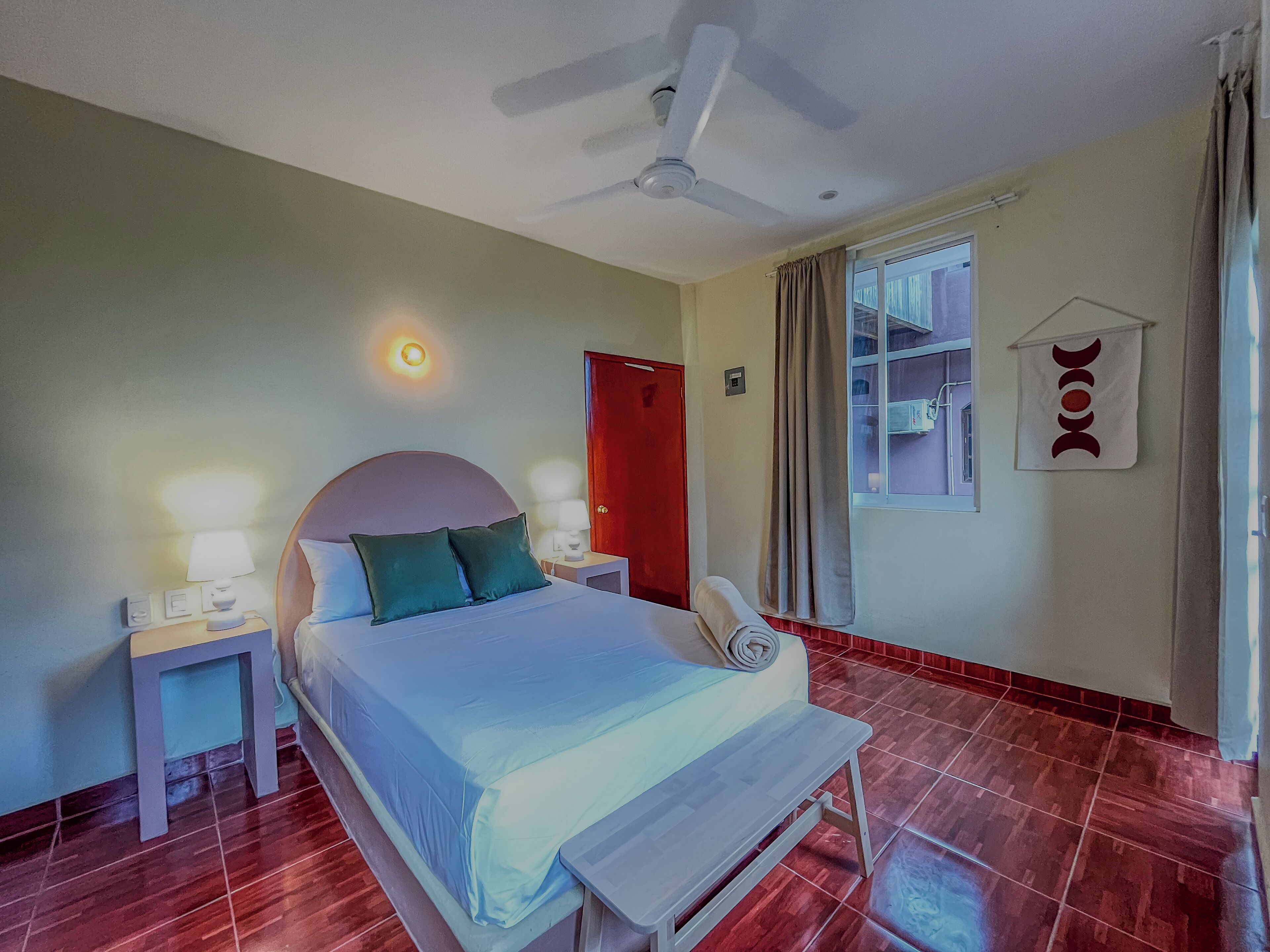 A hotel room with a bed, bedside tables, a ceiling fan, a window with curtains, and a wall-mounted decoration.