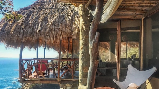Une villa en bord de mer dotée d’une terrasse en bois offrant une vue sur l’océan, d’un toit de chaume et d’une chaise blanche.