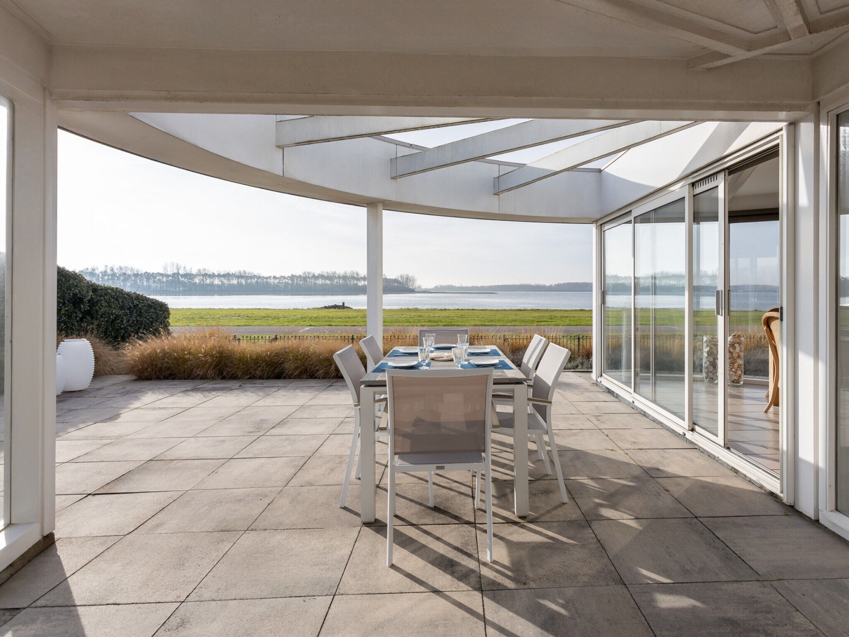 A covered patio with a table and chairs, large glass doors, and a view of a lake and fields.