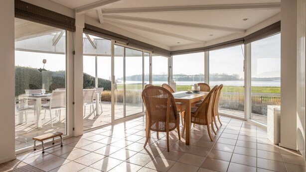 A spacious dining area with a large table and chairs, large windows offering a view of a lake and greenery, and a tiled floor. - Holiday Home by Lake Veerse Meer (Kamperland)