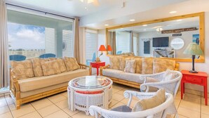 Ein Zimmer mit großem Fenster, Deckenventilator, Sofa, Sessel, kleinem Tisch und Spiegel.