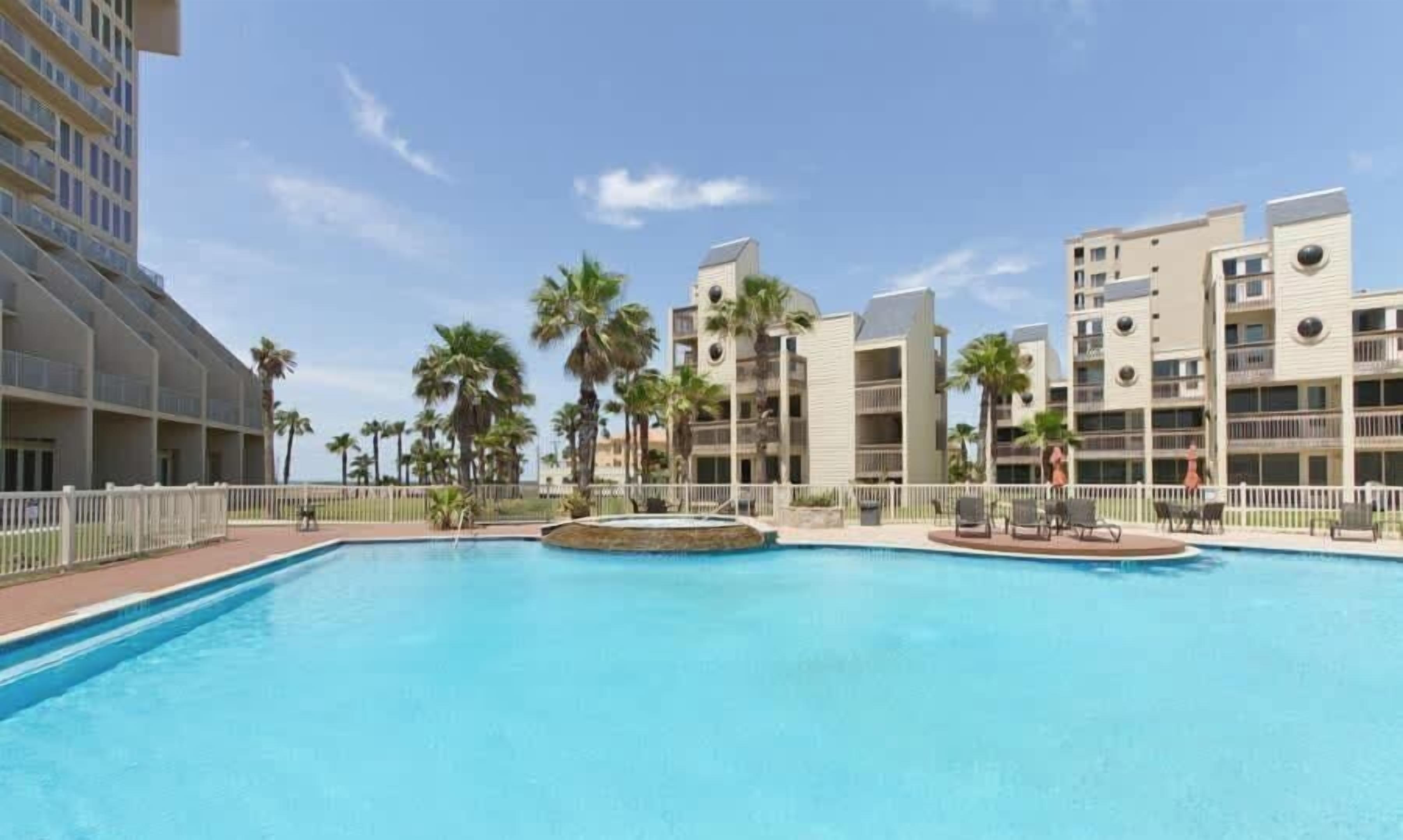 A swimming pool with a fountain, surrounded by palm trees and modern buildings.