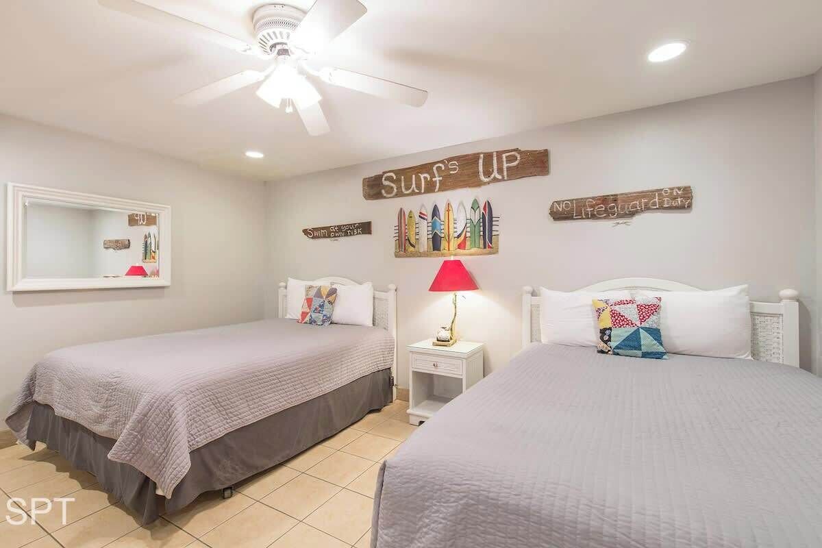 Un dormitorio con dos camas, un ventilador de techo y decoraciones en la pared, incluyendo tablas de surf y un cartel que dice 'Surf's Up'.