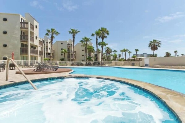 A swimming pool with a hot tub, surrounded by palm trees and a building with balconies.