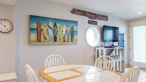 Ein Essbereich mit einem runden Tisch und Stühlen, einem Bild von Surfbrettern, einer Uhr, einem Schild mit der Aufschrift "South Padre Island, Texas" und einem Fernseher auf einem Schrank.