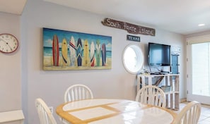 A dining area with a round table and chairs, a painting of surfboards, a clock, a sign reading "South Padre Island, Texas", and a television on a stand.