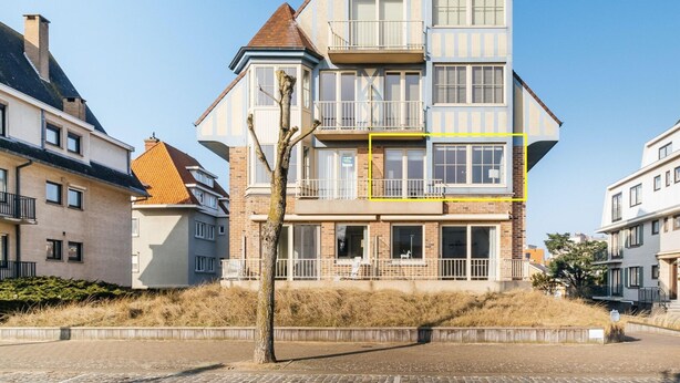 Un immeuble résidentiel à plusieurs étages, doté d’un balcon bien visible au deuxième étage, d’une façade en briques et, en arrière-plan, d’un ciel d’un bleu éclatant.