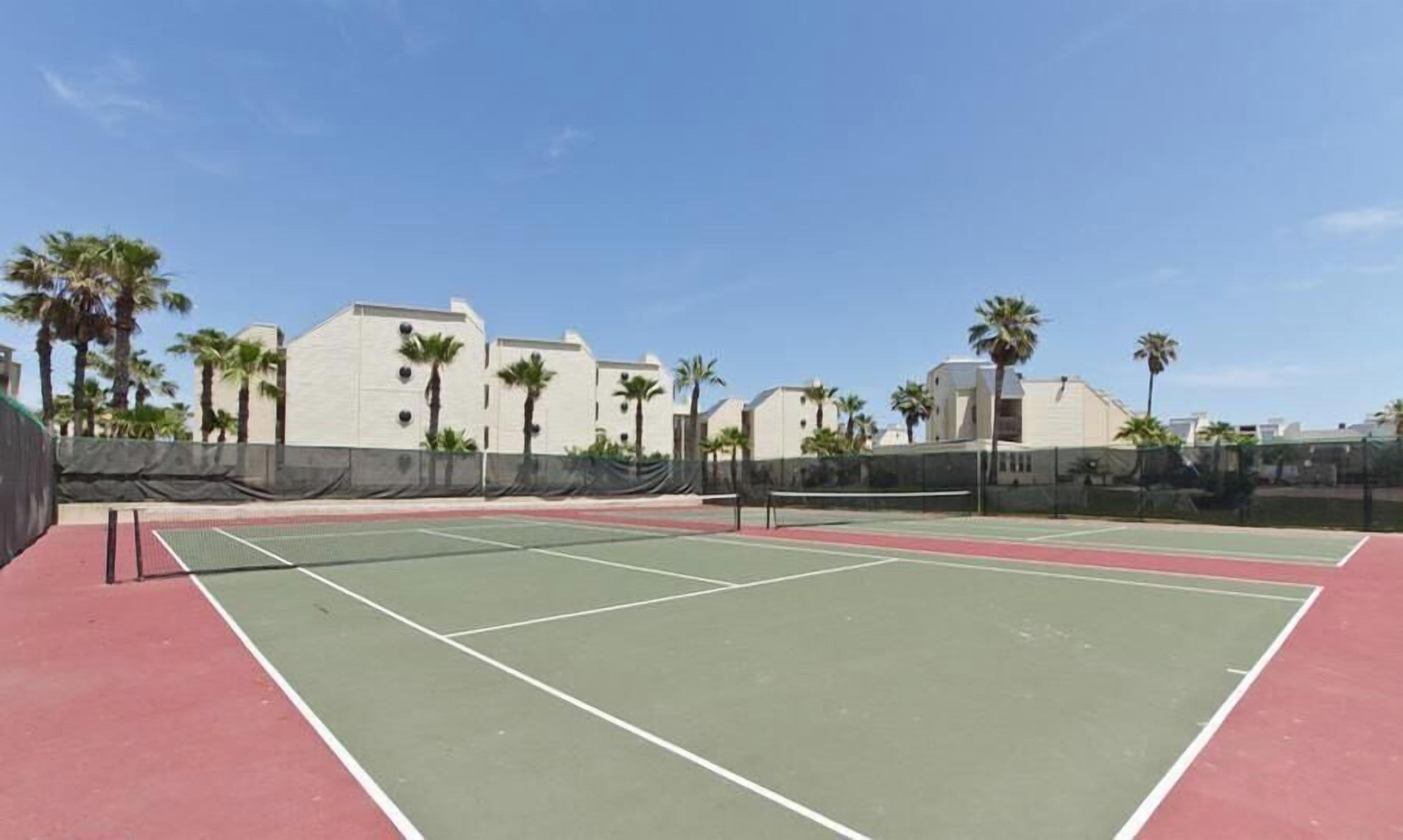 Un court de tennis à la surface rouge et verte, entouré de palmiers et d’immeubles.