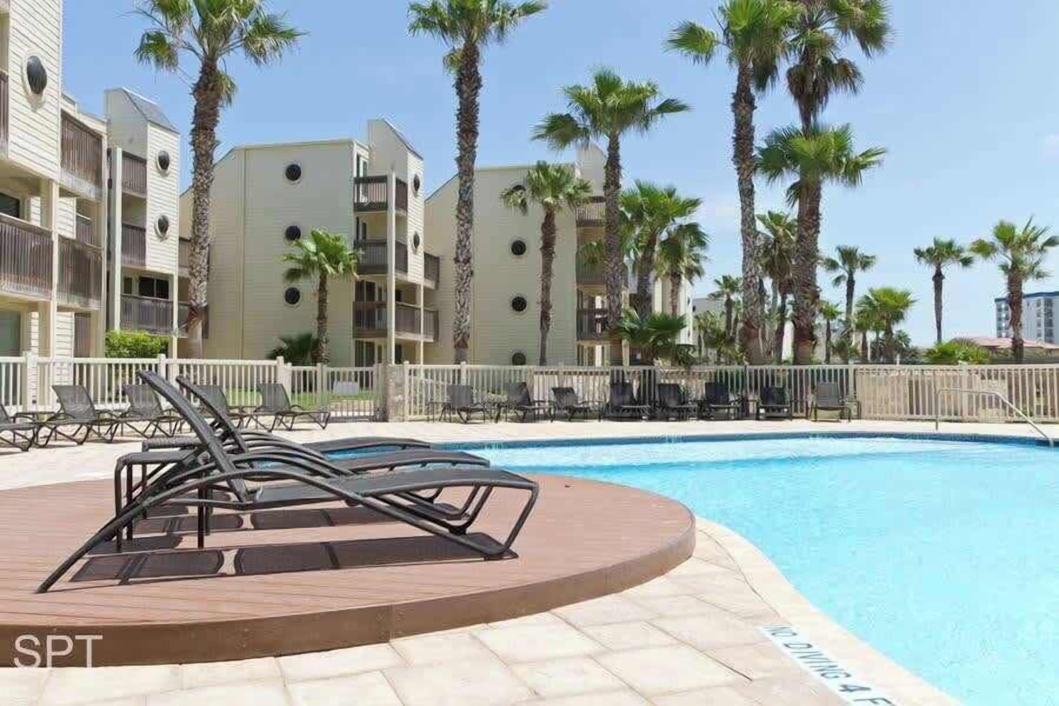 A swimming pool with lounge chairs, palm trees, and a multi-story building in the background.