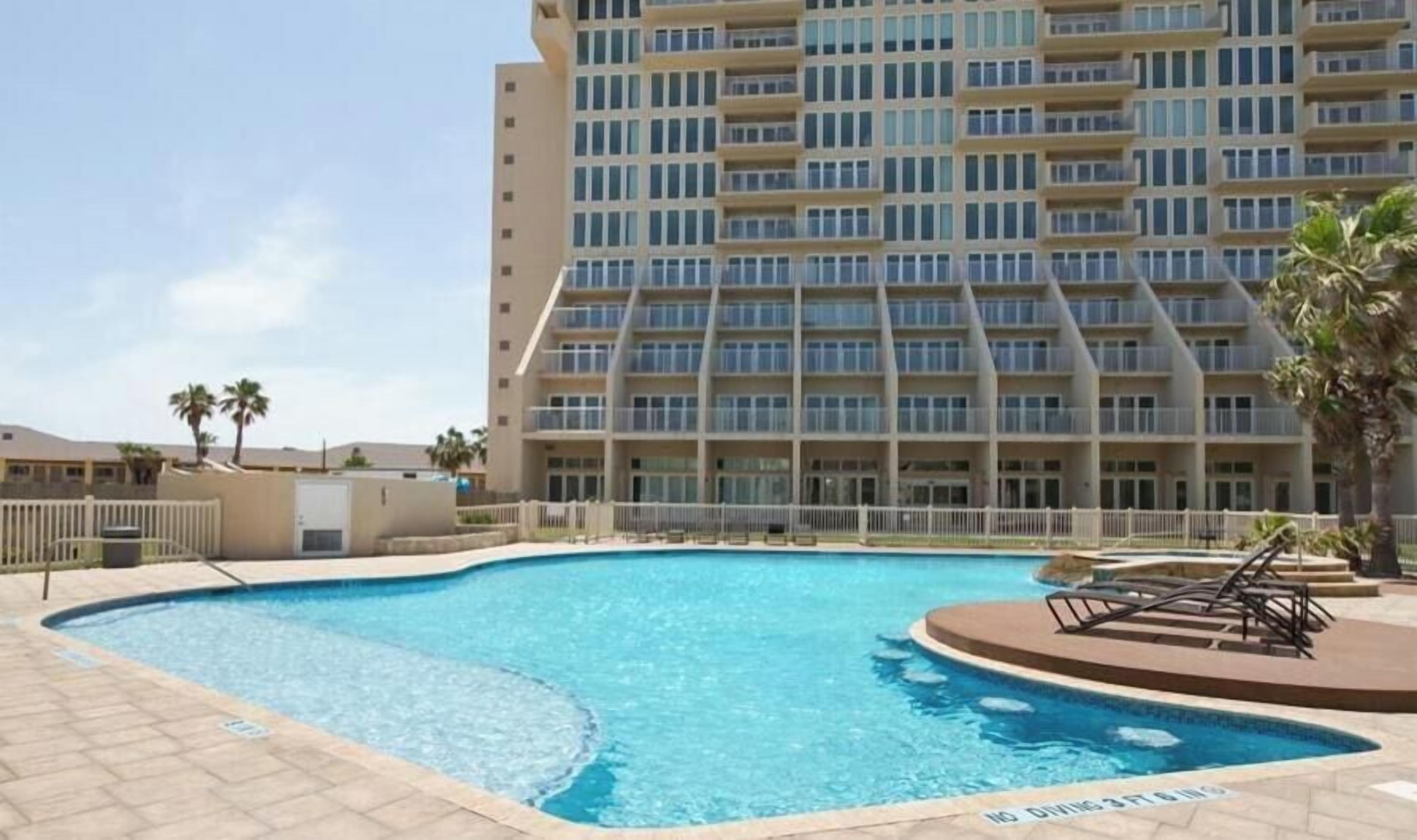 Una piscina con un cartel de 'No Diving', rodeada por una valla blanca, y un edificio de varios pisos con balcones al fondo.