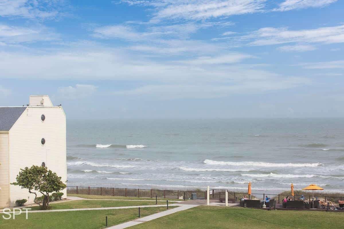 A coastal view with a building, a grassy area, and a seating area with umbrellas.