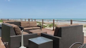 Een terras met rieten meubels en een glazen tafel met uitzicht op een strand.