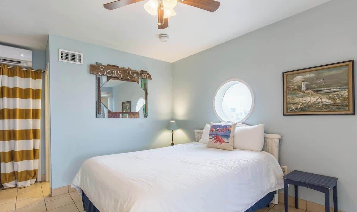 A bedroom with a bed, a ceiling fan, a window, a painting, and a sign on the wall.