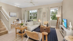 En moderne stue med sofa, sofabord og fjernsyn. Der er en trappe til venstre og et vindue med gardiner.