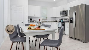 A modern kitchen with white cabinets, a stainless steel refrigerator, a microwave, and a dining area with a white table and grey chairs. - Seas the Day Beach Wine Dine Relax (Naples)