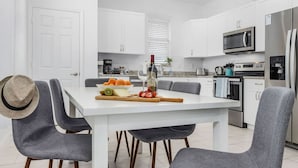 A modern kitchen with white cabinets, a stainless steel refrigerator, and a dining area with grey chairs and a white table. - Seas the Day Beach Wine Dine Relax (Naples)